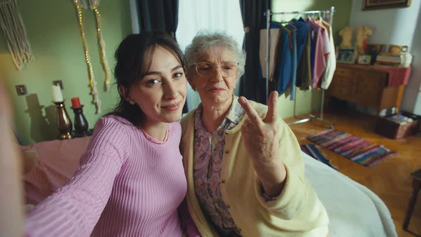 A loving granddaughter and her grandmother make a fun selfie video together, smiling, kissing, and making heart signs for the camera