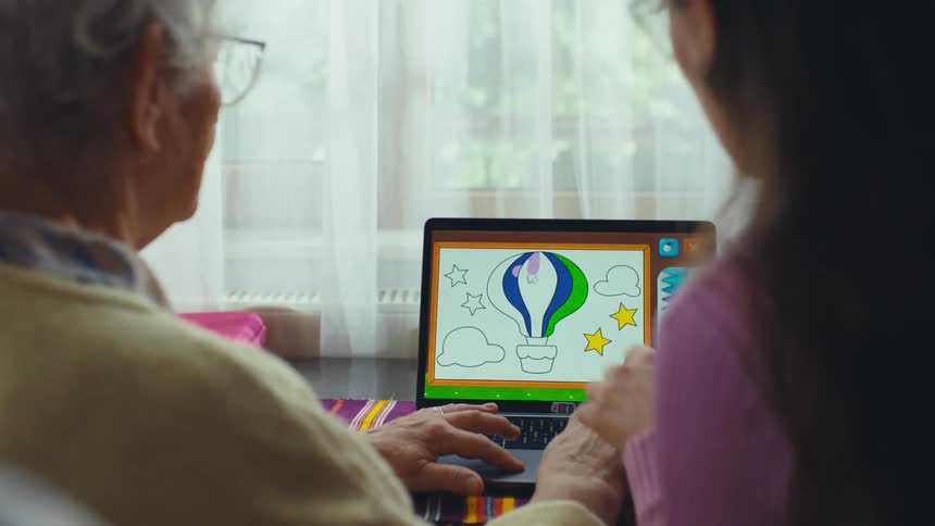 From an over-the-shoulder perspective, a young girl teaches her grandmother how to use a laptop to color in a digital coloring book