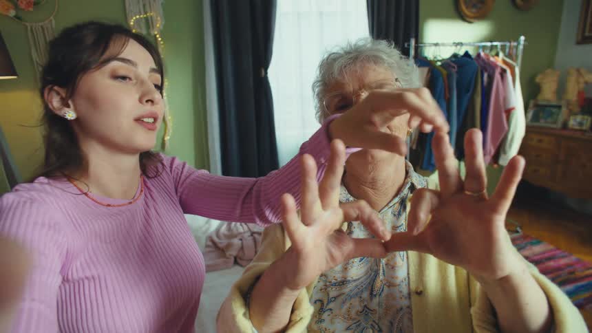 A loving granddaughter teaches her elderly grandmother how to make a heart gesture with her hands for a fun selfie video in a cozy bedroom