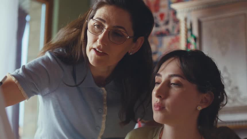 A senior artist guides her young student through a painting, sharing her wisdom in a sunlit studio