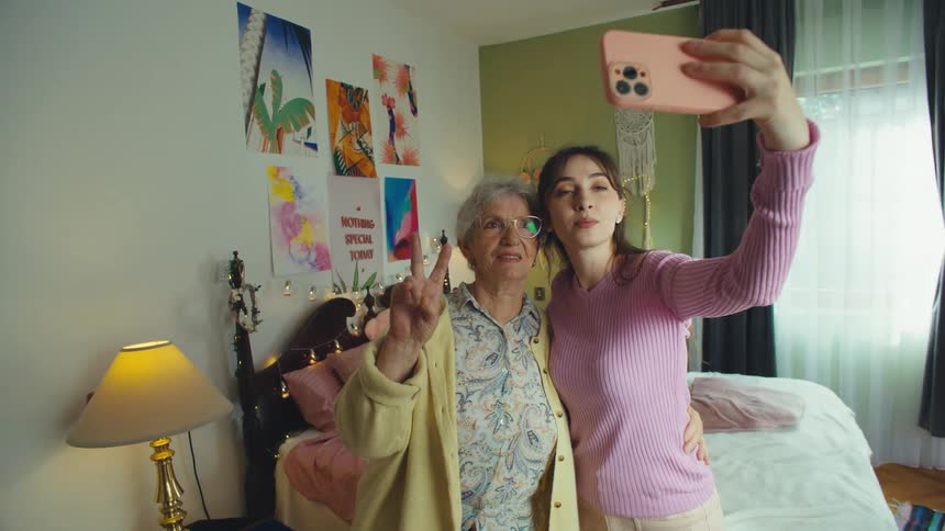 A granddaughter and her grandmother have fun making a video for social media, dancing and posing with peace signs in a bedroom