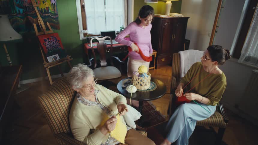Three generations of women, a grandmother, mother, and daughter, share a joyful moment knitting together in their cozy living room