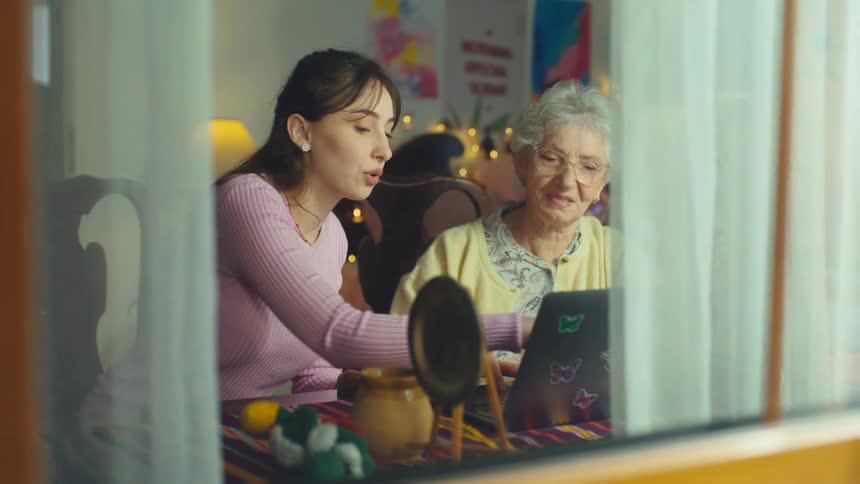 A young woman patiently teaches her elderly grandmother how to use a laptop, viewed from outside through a window