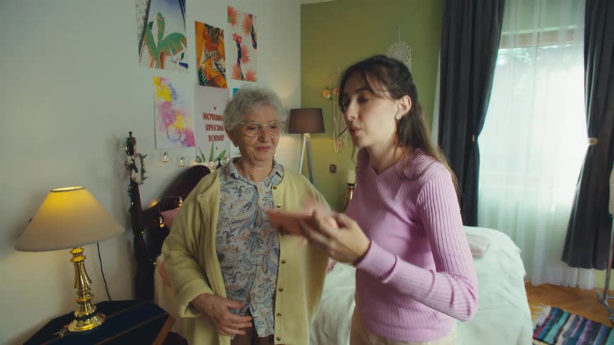 A happy granddaughter and her elderly grandmother take fun selfies together with a smartphone in a colorful bedroom