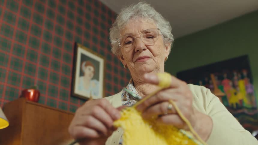 A serene elderly woman with glasses carefully knits with bright yellow yarn in a cozy, vintage-style living room