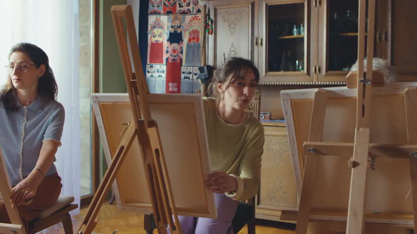 A young female art teacher leads a painting class for two adult women in a cozy, sunlit home studio