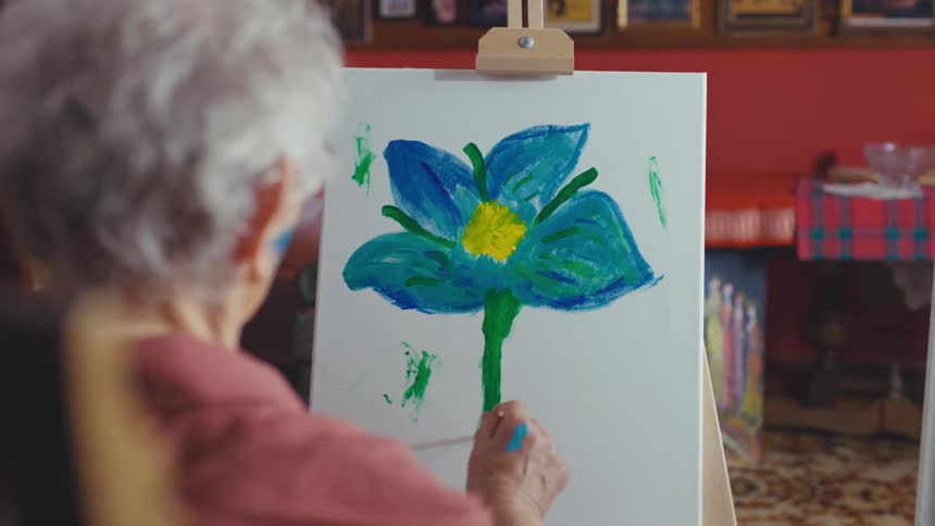 An elderly woman with white hair paints a vibrant blue and yellow flower on a canvas inside her home