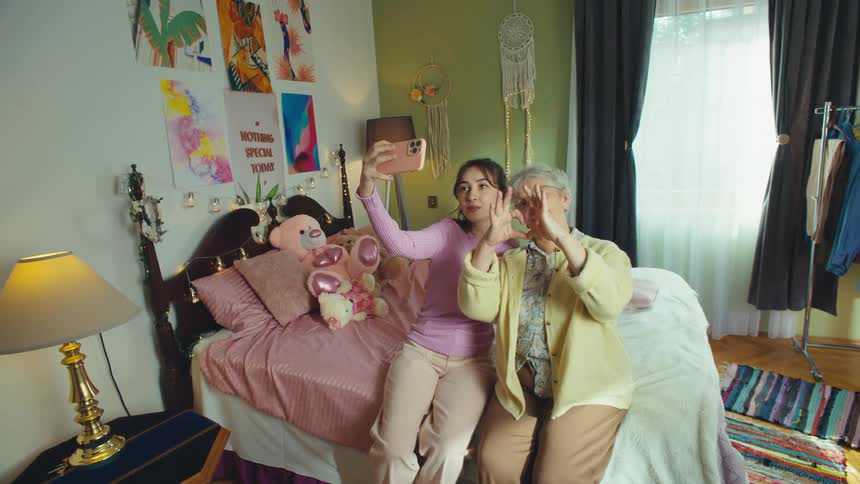 A young woman and her grandmother take a fun selfie together, making heart shapes and peace signs in a cozy bedroom