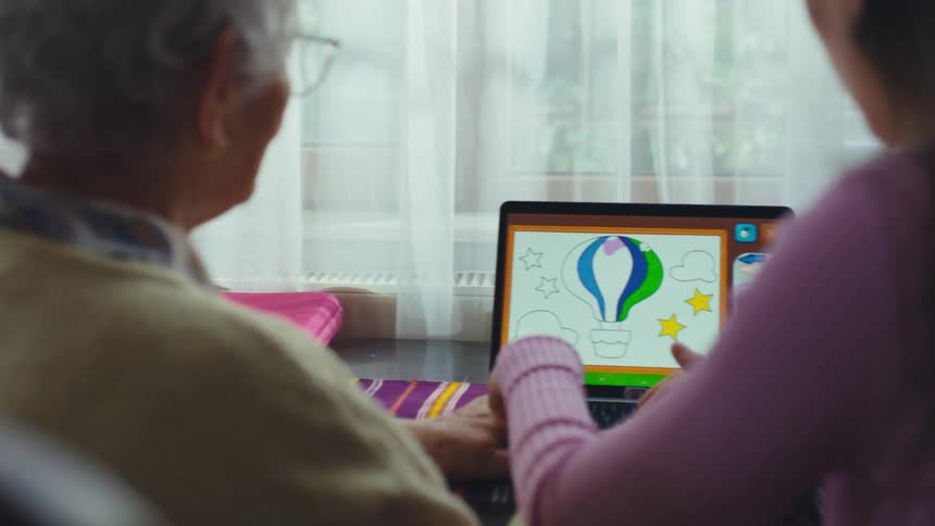 An elderly grandmother smiles as her granddaughter teaches her how to color on a laptop using a digital coloring book application