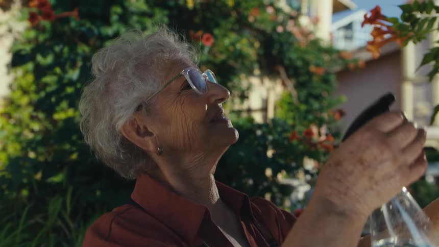A serene elderly woman in an apron lovingly tends to her vibrant flowering vine, gently spraying it with water in her sun-drenched garden
