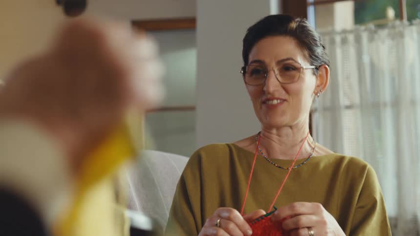 A woman with glasses knits with red yarn and laughs while talking with a friend in a cozy, sunlit room