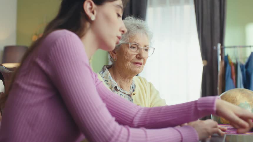 A young woman patiently helps her elderly grandmother navigate a laptop computer in a warm, cozy room