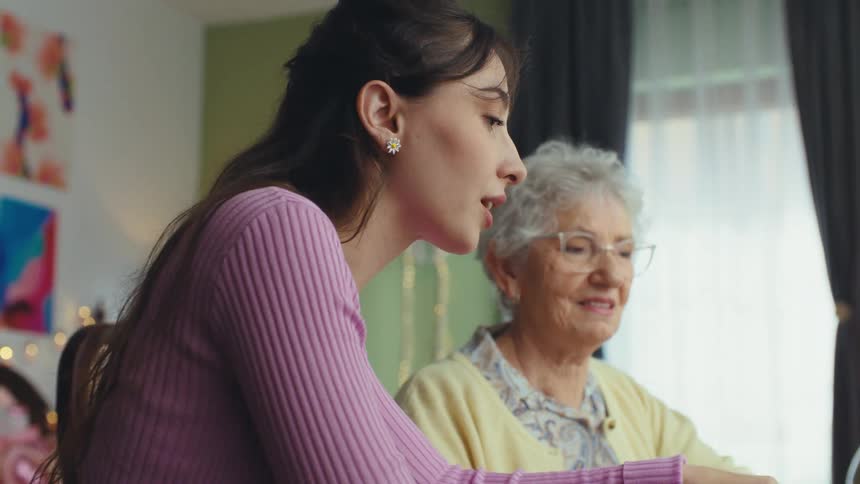 A caring granddaughter sits with her elderly grandmother, patiently teaching her how to use a laptop in a cozy home environment