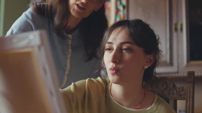 An art teacher guides her female student as she paints on a canvas, offering advice and encouragement in a warm, sunlit studio