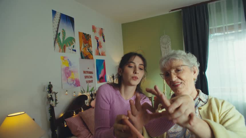 A granddaughter and her grandmother have fun taking a selfie video in the bedroom, making a heart shape with their hands