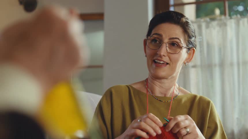 A middle-aged woman knits and talks with an elderly woman, with the camera focus shifting to the older woman's hands knitting