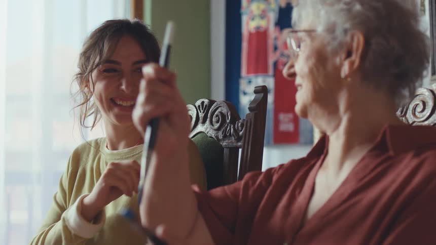 A granddaughter playfully teases her artistic grandmother with a paintbrush as they laugh and bond during a painting session at home