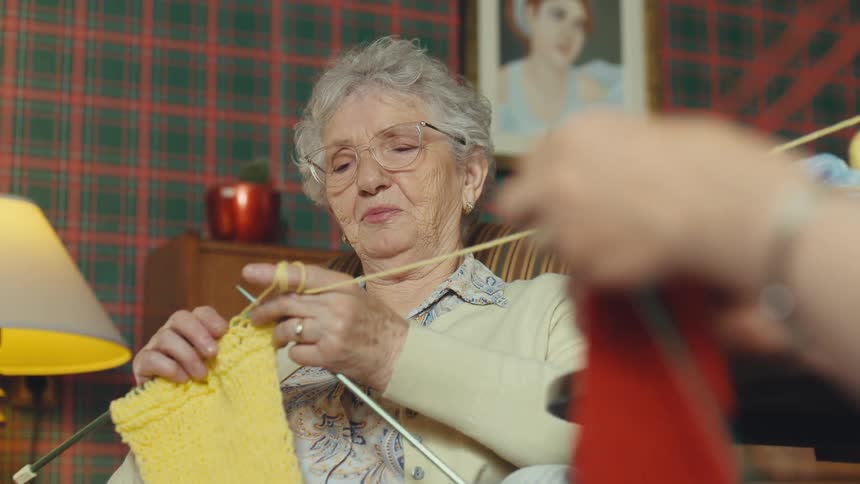 A calm elderly woman with glasses knits with yellow yarn while sitting in a cozy, vintage-style living room