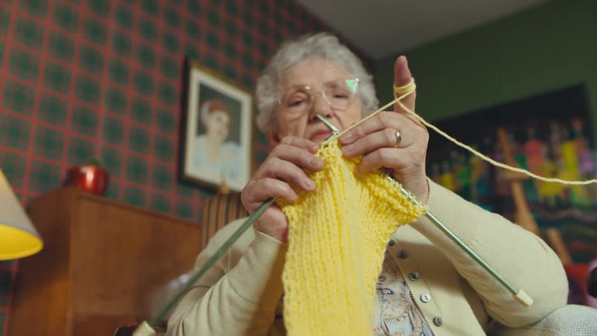 An elderly woman with gray hair and glasses carefully knits with yellow yarn in her cozy, retro-style living room