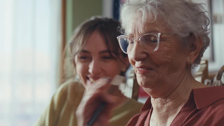 A cheerful young woman affectionately watches her elderly grandmother paint, sharing smiles and a warm embrace during a creative afternoon