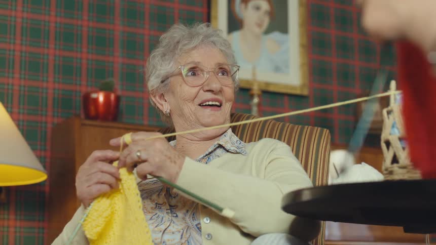 A cheerful elderly woman with glasses laughs and smiles while knitting with yellow yarn in her cozy living room