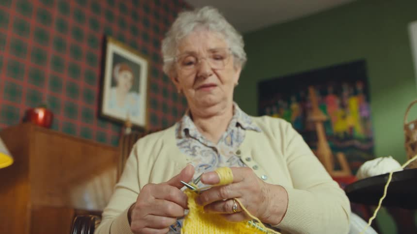 Grandmother teaches her granddaughter how to knit with yellow yarn in a cozy, vintage-style living room