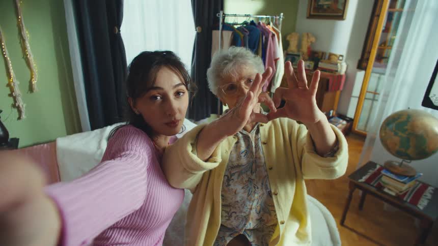 A young woman and her grandmother take a fun selfie video, making peace signs and heart shapes with their hands in a cozy bedroom