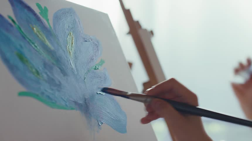 Close-up of a young artist painting a blue flower on a canvas during a lesson with her instructor