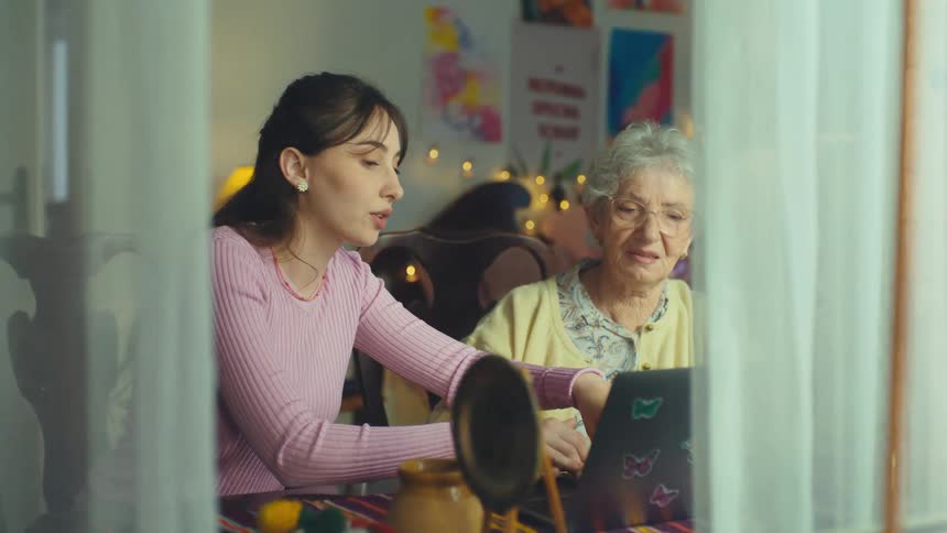 A young woman patiently teaches her elderly grandmother how to use a laptop computer in a cozy room, as seen through a window