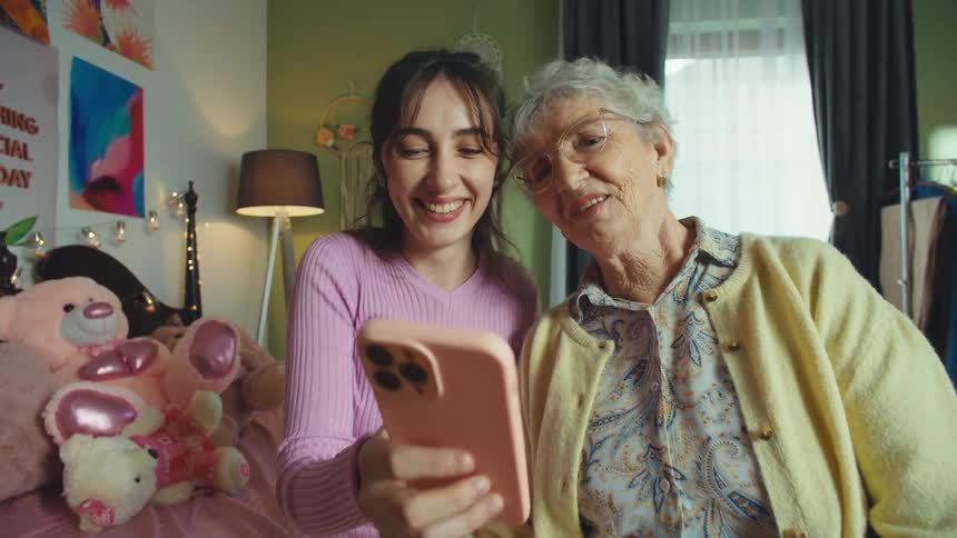 A smiling granddaughter shows her grandmother something on her smartphone as they sit together in a cozy bedroom