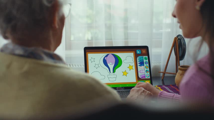 A young woman and her elderly grandmother spend quality time together, using a laptop to color in a digital drawing of a hot air balloon