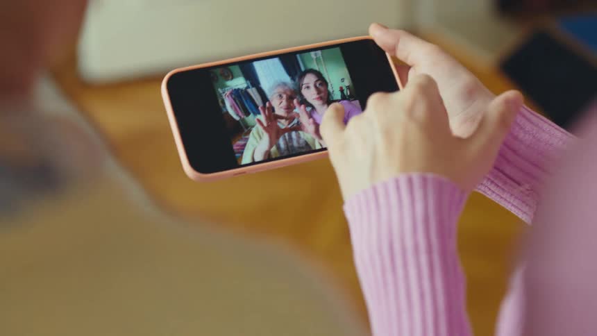 Close-up over the shoulder view of a woman scrolling through a gallery of selfies with her elderly grandmother on a smartphone