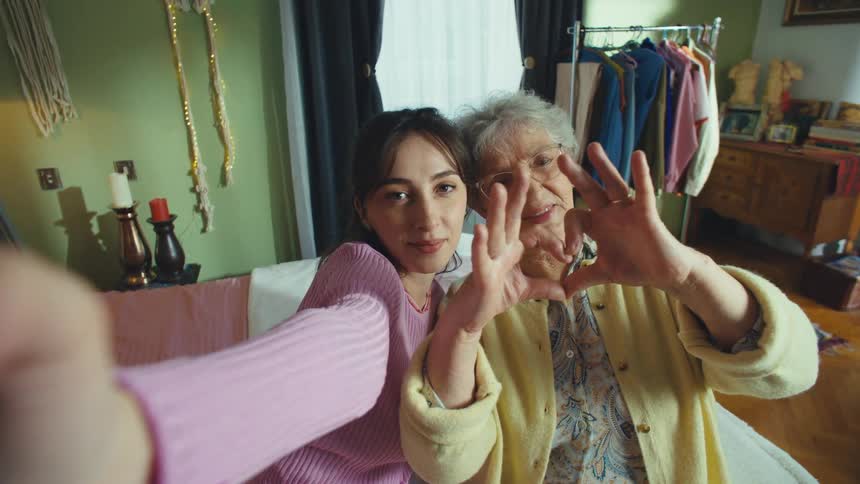 A young woman and her grandmother film a sweet selfie video, smiling and making a heart shape with their hands for the camera