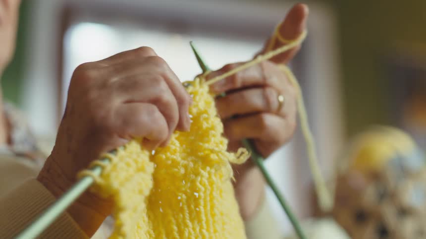 A close-up shot of an elderly woman's hands skillfully knitting with bright yellow yarn in a cozy indoor setting