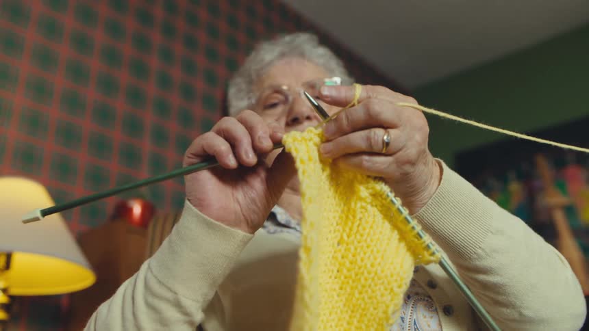 A slow dolly-in shot focuses on an elderly woman knitting with yellow yarn in her cozy, vintage-style living room
