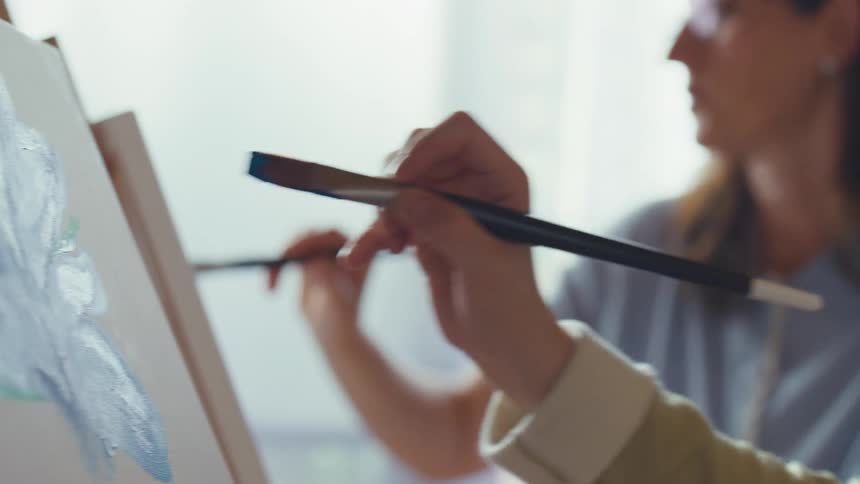 Close-up of two women artists painting side-by-side in a bright and calm studio, focusing on mixing paint and applying brushstrokes to a canvas