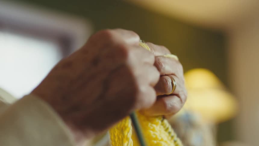 Closeup shot pans from an elderly woman's face down to her hands as she carefully knits with yellow yarn