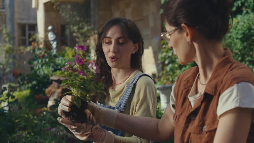 A mother and her teenage daughter share a warm moment while gardening together, passing a vibrant flowering plant in their sunny backyard