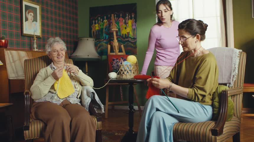 Three generations of women share a heartwarming moment as the mother and grandmother knit a scarf for the daughter in their cozy living room
