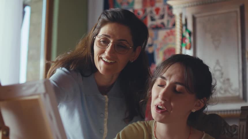 A female art teacher gives friendly advice to her student during a painting class in a sunlit studio