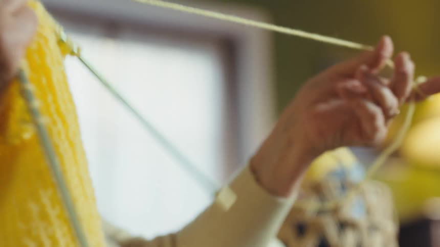 A slow-motion close-up of an elderly woman's hands skillfully knitting with bright yellow yarn in a cozy room