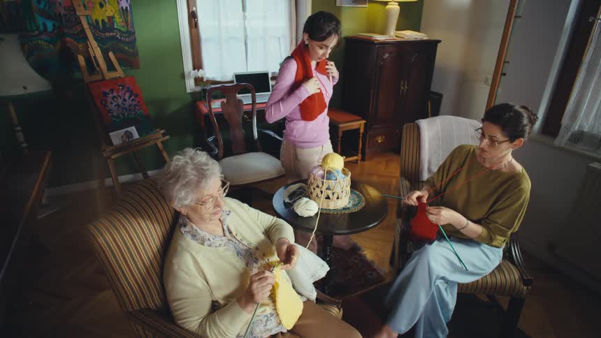 A young woman comically tries on a knitted scarf her mother is making while her grandmother watches and knits, sharing a happy family moment