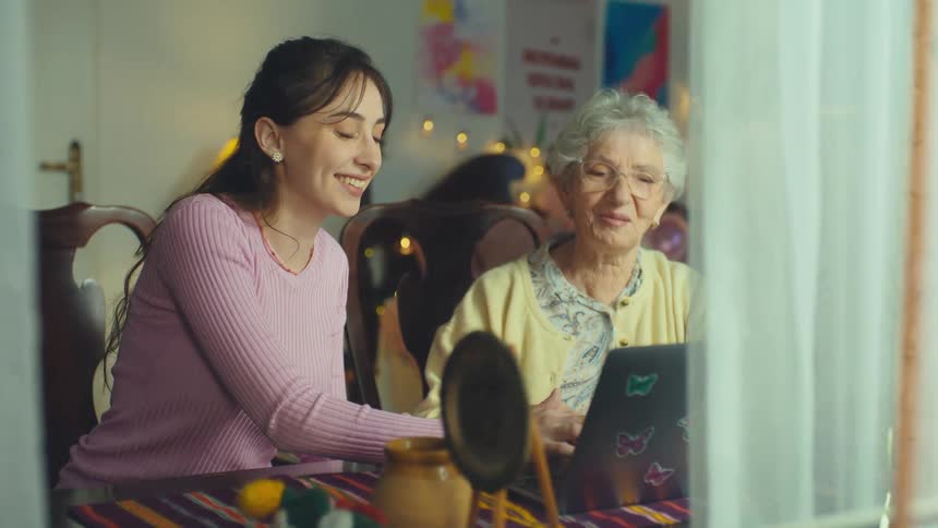 A caring young woman patiently teaches her elderly grandmother how to use a laptop, viewed from outside their cozy home