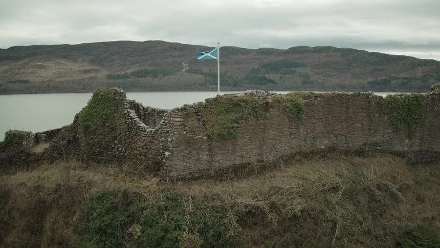 A Scottish flag waves in the wind above the ancient stone ruins of a castle on the shores of a Scottish loch