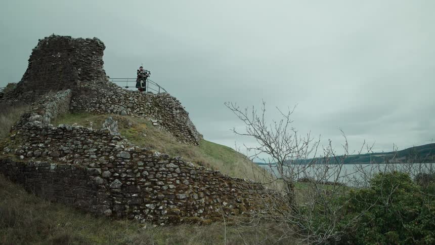 A solitary Scottish bagpiper in a traditional kilt plays atop the ancient ruins of Urquhart Castle as the camera reveals the iconic Loch Ness