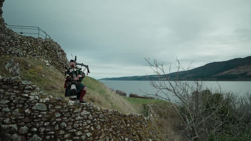 A crane shot reveals a Scottish bagpiper in a traditional kilt playing on the stone walls of Urquhart Castle overlooking Loch Ness