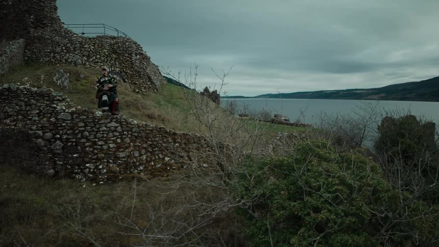 An aerial shot flies past a bagpiper on the ancient ruins of Urquhart Castle to reveal the historic site on the banks of Loch Ness, Scotland