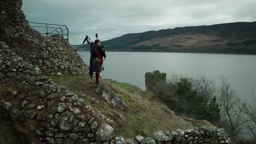 Aerial shot reveals a bagpiper in a traditional kilt playing at the ruins of Urquhart Castle on the shores of Loch Ness in Scotland