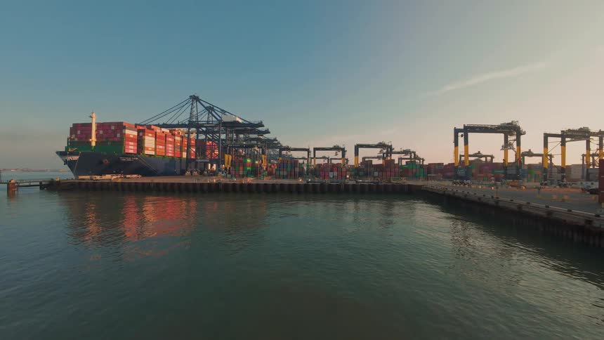 A large container ship loaded with cargo is docked at a busy shipping port as cranes operate along the pier