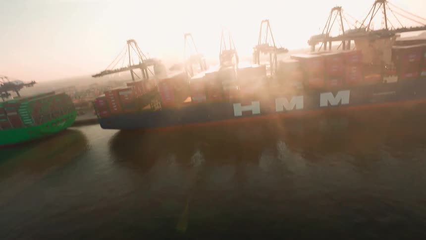 A dramatic aerial reveal shows a massive container ship docked at an industrial port with cranes during a hazy sunrise